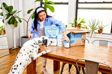 A woman unpacking a Spot & Tango delivery box of UnKibble bags and fresh food onto a wooden table while a Dalmatian watches
